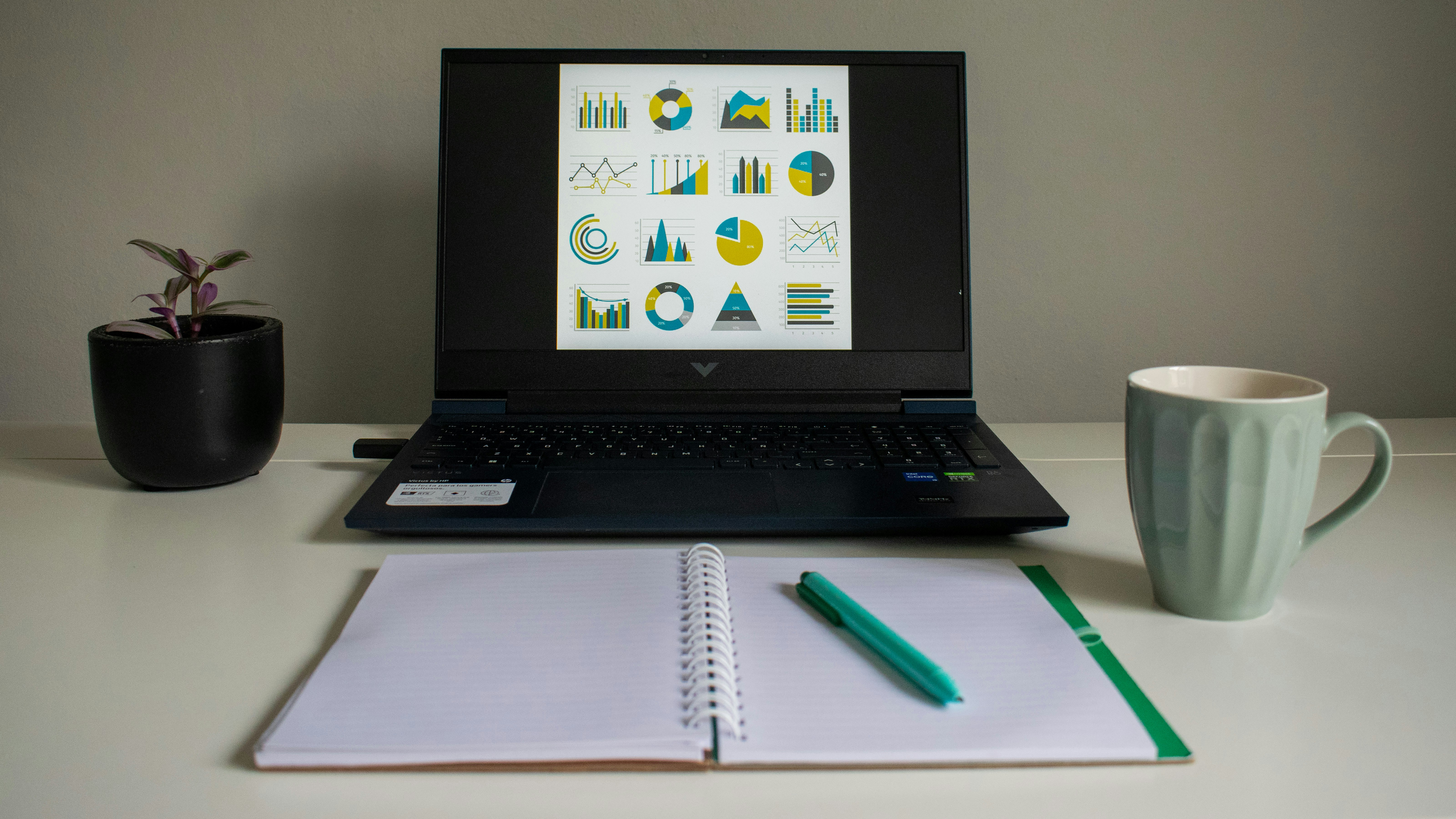 Laptop displaying charts beside a notebook and mug